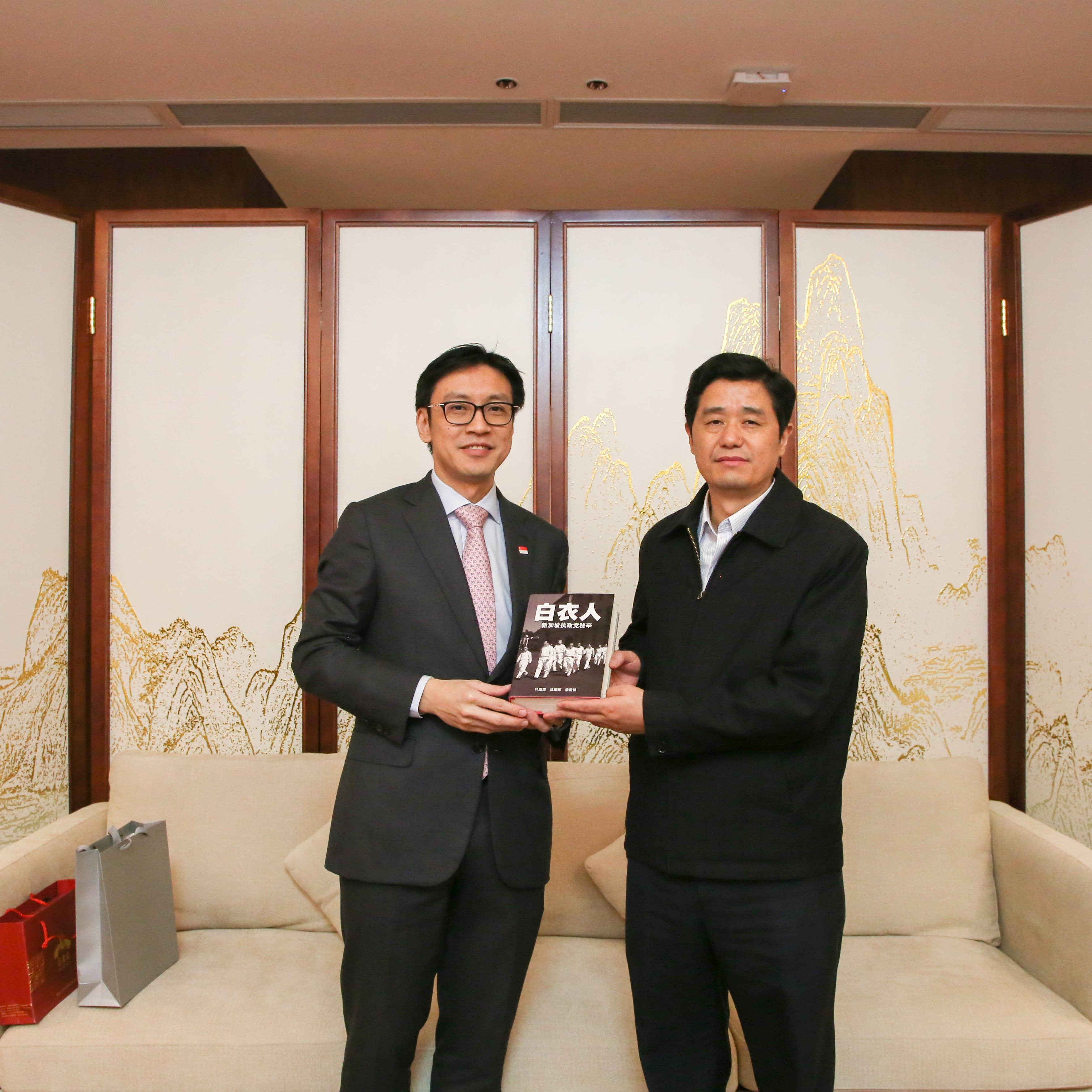 Two men holding book "白衣人" (The Whites) with black and white photograph on cover, standing indoors.